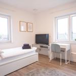 Schlafzimmer mit Bett, Schreibtisch, Fernseher und zwei Fenstern.