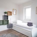 Ein Schlafzimmer mit weißem Bett, Holzboden und Wandregalen. An der Wand hängen Bilder.