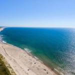 Ausblick auf einen sandigen Strand mit türkisfarbenem Meer und grünen Dünen.