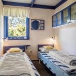 Zwei Einzelbetten mit blauen Kopfenden und gelben Kissen. Fenster mit blauem Rahmen und Bücherregal.