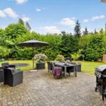 Terrasse mit Grill, Tisch, Stühlen und Sonnenschirm. Grünflächen und Bäume im Hintergrund.