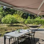 Terrasse mit Tisch, Stühlen und Hängemöbel unter Markise. Grüner Garten im Hintergrund.