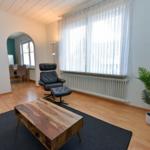 Wohnzimmer mit Sitzgelegenheit, Holztisch und Pflanze. Durchgang zur Küche.