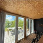 Raum mit Holzdecke und großen Fenstern zum Balkon mit Tisch und Stühlen.