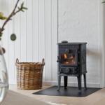 Black stove with burning fire stands on wooden floor next to basket and plant.