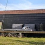 Outdoor bench with cushions and blanket in front of dark wooden house with chimney and tiled roof.