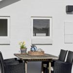 Terrace with table, chairs, plant, and kettle in front of white wall with windows.