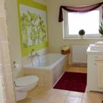 Badezimmer mit Badewanne, Waschbecken und Toilette. Wandbild und Fenster mit Vorhang.