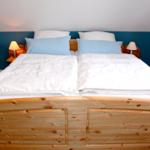 Doppelbett mit Holzrahmen, weißen Bettwäsche und blauen Kissen.