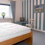 Ein Schlafzimmer mit Bett, Schreibtisch, Stuhl und Streifenwand. Fenster mit Vorhängen und Pflanze.