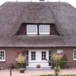 Haus mit strohgedecktem Dach, weißen Fenstern und Eingangstür. Rote Blumen vor dem Eingang.