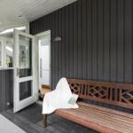 Holzbank mit weißer Decke und Buch auf Terrasse vor Haus mit dunkler Fassade.