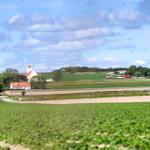 Weitläufige Landschaft mit Feldern, einem Dorf und einer Kirche unter blauem Himmel.
