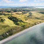 Aerielansicht einer Küstenlandschaft mit Grashügeln, Wald und Strand. Ein Haus ist am Ufer sichtbar.