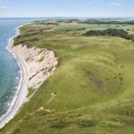 Aerial view of coastal cliffs, green hills, and sea under clear sky.