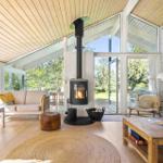 Wohnraum mit Kamin, Sofa und Holzboden. Große Fenster mit Blick auf Garten.