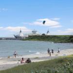 Strand mit Menschen, Segelboot und Kitesurfer im Hintergrund.