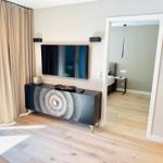 Wohnbereich mit TV, Sideboard und Blick in Schlafzimmer. Holzboden und Fenster mit Vorhängen.