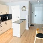 Küche mit Arbeitsplatte, Tisch und Stühle. Wanduhr und Holzboden sichtbar.