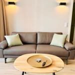 Ein braunes Sofa mit zwei Kissen steht vor einem Fenster. Davor ein runder Tisch mit einem goldenen Tablett.