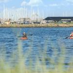 Zwei Personen paddeln in Kanus auf dem Wasser vor einem Hafen mit Booten und einem Gebäude.