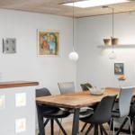 Esszimmer mit Holztisch, Stühlen und Wandbildern. Deckenleuchten und Regal mit Pflanzen.