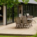 Terrasse mit Holztisch und Stühlen vor einem Haus mit Glaswänden.