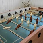 Fussballtisch mit Holzrahmen und Metallstangen. Hintergrund: weiße Wand und Pflanze.