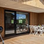 Terrasse mit Grill, weißem Tisch und Stühlen, Holzwand und Glastüren.