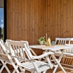 Terrasse mit weißem Tisch und Stühlen, Holzwand im Hintergrund, Sonne scheint.