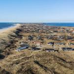 Aerielansicht von Strandhäusern auf Dünen mit Windkraftanlagen am Horizont.