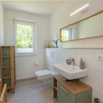 Badezimmer mit Waschbecken, Spiegel, Toilette und Fenster mit Blick auf Grün.