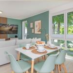 Wohn- und Essbereich mit Sofa, Tisch und Stühlen. Große Fenster mit Blick auf Bäume.