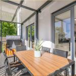 Terrasse mit Holztisch, Stühlen und Blick auf grüne Bäume. Durchgehende Glaswände führen ins Haus.