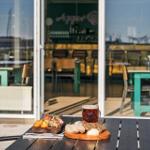Außenbereich mit Tisch, Getränken und Speisen. Im Hintergrund Restaurant mit Glasfront.