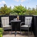 Terrasse mit zwei Stühlen und Tisch, umgeben von grüner Vegetation und dunklem Holzzaun.