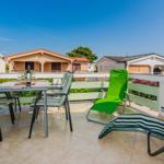 Terrace with table, chairs, and loungers. Background: houses and flowers.
