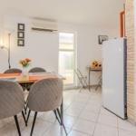 Dining area with table, chairs, and refrigerator. Window and plants visible.