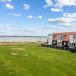 Grüner Rasenbereich mit Blick auf das Wasser und moderne Häuser mit roten Dächern.