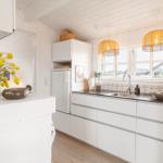 Küche mit weißen Schränken, Arbeitsplatte und Fenster. Hängelampen und Blumen sind sichtbar.