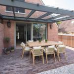 Terrasse mit Holztisch, Stühlen und Glasdach. Roter Backsteinbau und Pflanzen sichtbar.