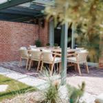 Terrasse mit Holztisch, Stühlen und Pflanzen unter Glasdach.
