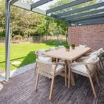 Terrasse mit Tisch und Stühlen unter Pavillon. Hintergrund: grüner Garten und Backsteinmauer.
