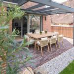 Terrasse mit Holztisch und Stühlen unter Glasdach. Hintergrund: Haus mit rotem Ziegeldach.