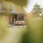 Haus mit Terrasse und Garten. Holztisch mit Stühlen. Blick durch Blätter.