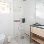 Bad mit Dusche, Waschbecken und Toilette. Glaswand, Holzunterschrank und Fenster.