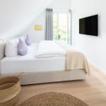 Schlafzimmer mit Bett, TV und Fenster. Holzboden und Wandlampe sichtbar.