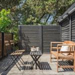 Terrasse mit Holzbank, Tisch und Stühlen. Umgeben von Holzwänden und Bäumen.