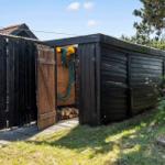Holzschuppen mit offenem Tor, darin Schlauch und Holzscheite, umgeben von Grünflächen und Zaun.
