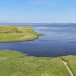 Weitläufiger Blick auf grüne Wiesen und ruhiges Wasser unter blauem Himmel.